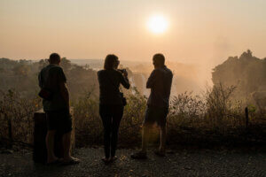 three travellers standing by the edge of the Batoka gorge into which the mighty Zambezi River plunges as the majestic Victoria Falls. The spray from the falls making the sunset more enchanting